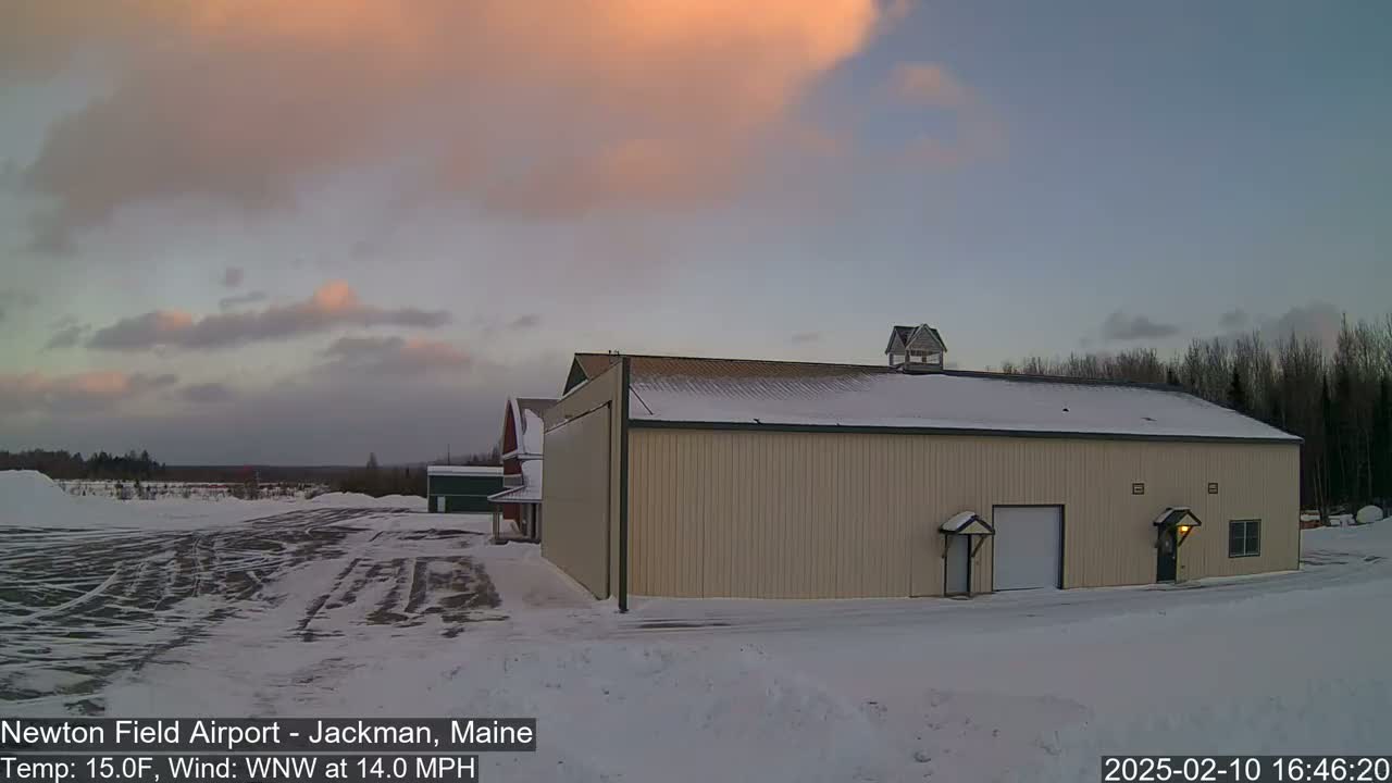 A large tan hangar sits in a snow-covered airfield under a twilight sky with pale pink clouds and a 14 mph wind.