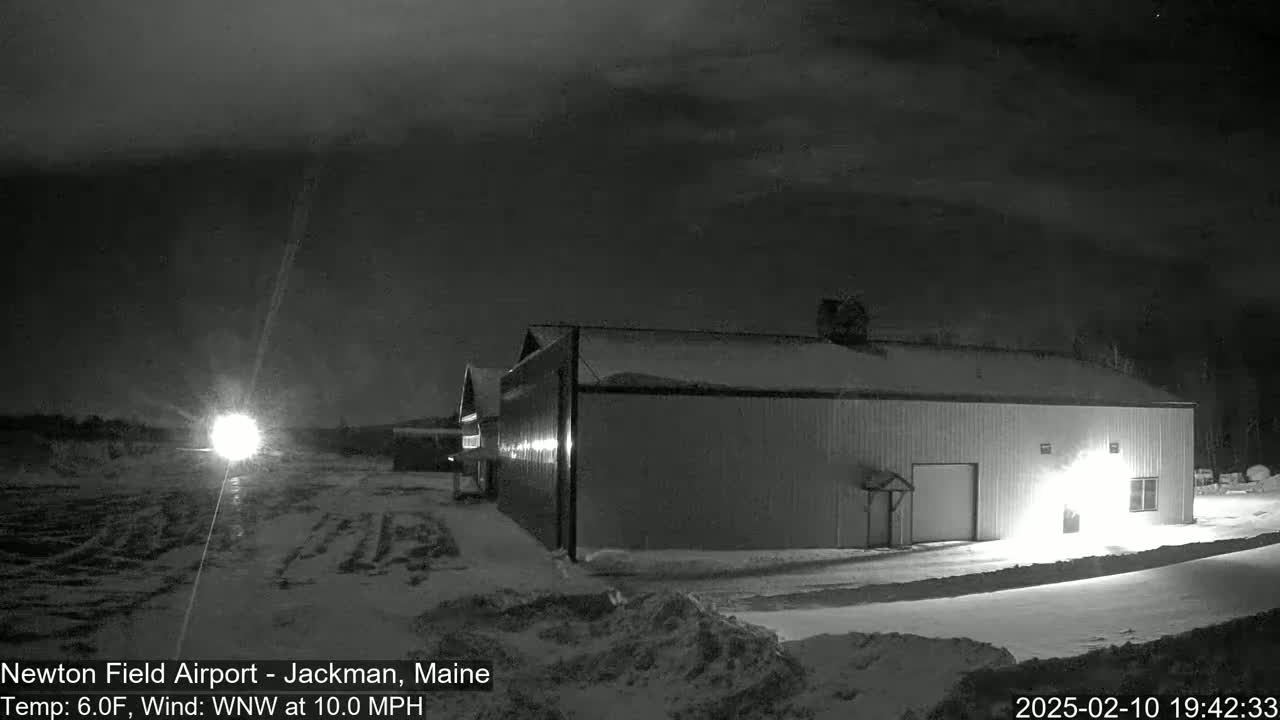A large, dark hangar sits in a snow-covered field at night under a cloudy sky, with a bright light visible in the distance.