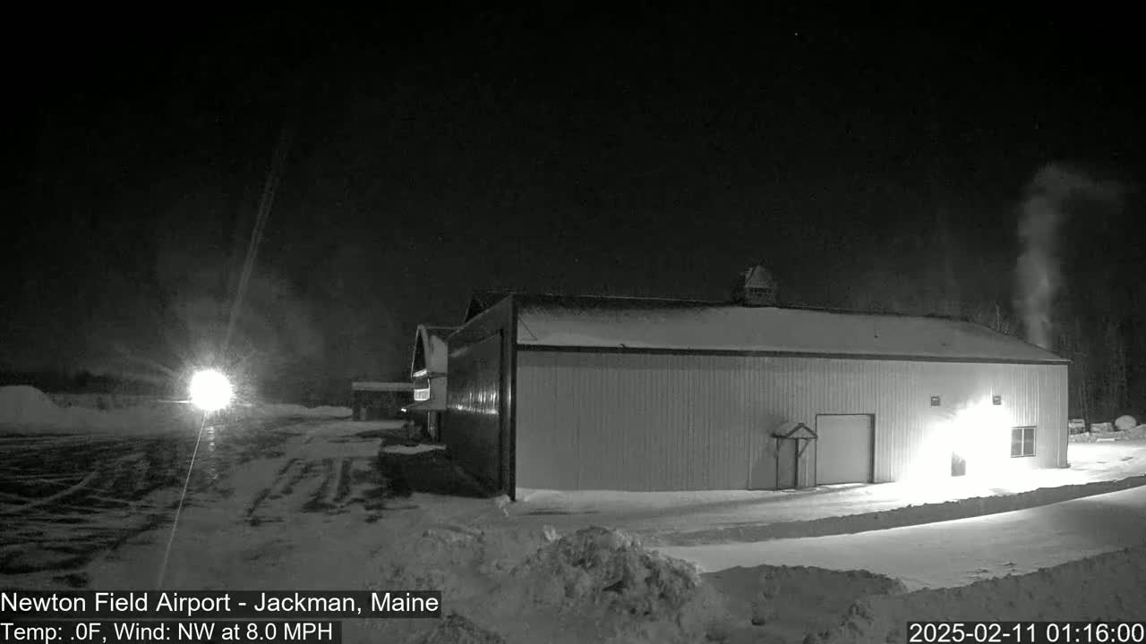 A long, snow-covered building is illuminated at night under a dark, clear sky with a northwest wind of 8 mph and a temperature of 0°F.