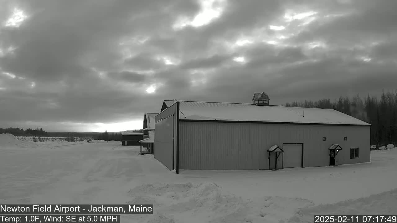 A large, snow-covered hangar-like building stands in a snowy landscape under a mostly cloudy sky, with a slight hint of sunrise or sunset.