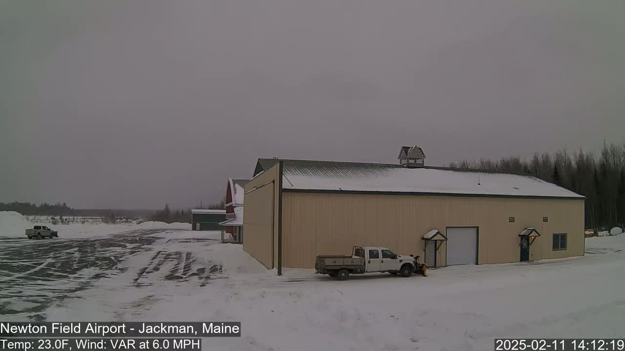 A tan hangar and a red barn sit on a snow-covered airfield under an overcast sky.