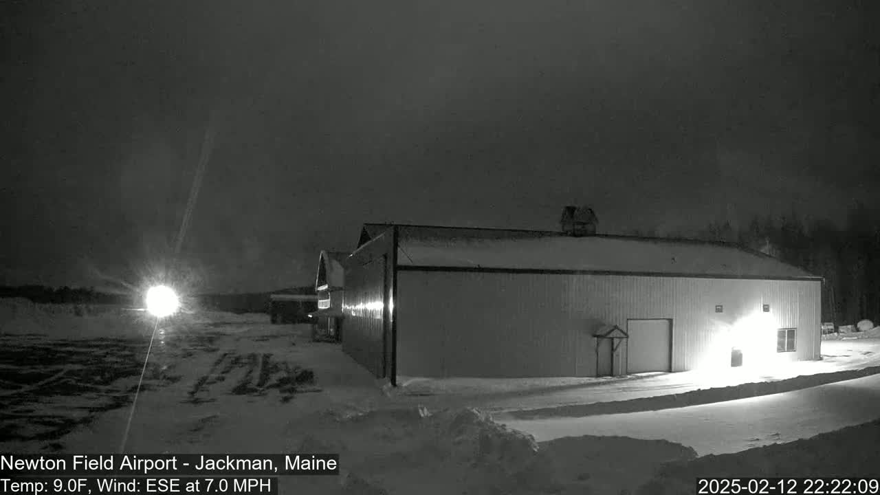 A large, snow-covered hangar is illuminated at night under a dark, overcast sky with a light source visible in the distance.