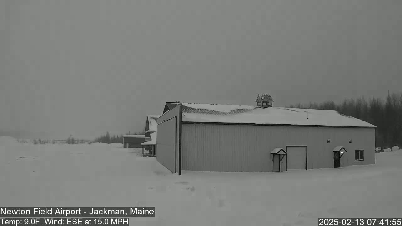 A snow-covered hangar and other buildings are visible in a snowy landscape under an overcast sky with a wind of 15 mph from the east-southeast.