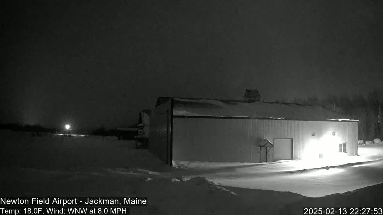 A snow-covered landscape at night shows a large, single-story building illuminated by exterior lights, under a dark, clear sky with a wind of 8.0 mph from the west-northwest.