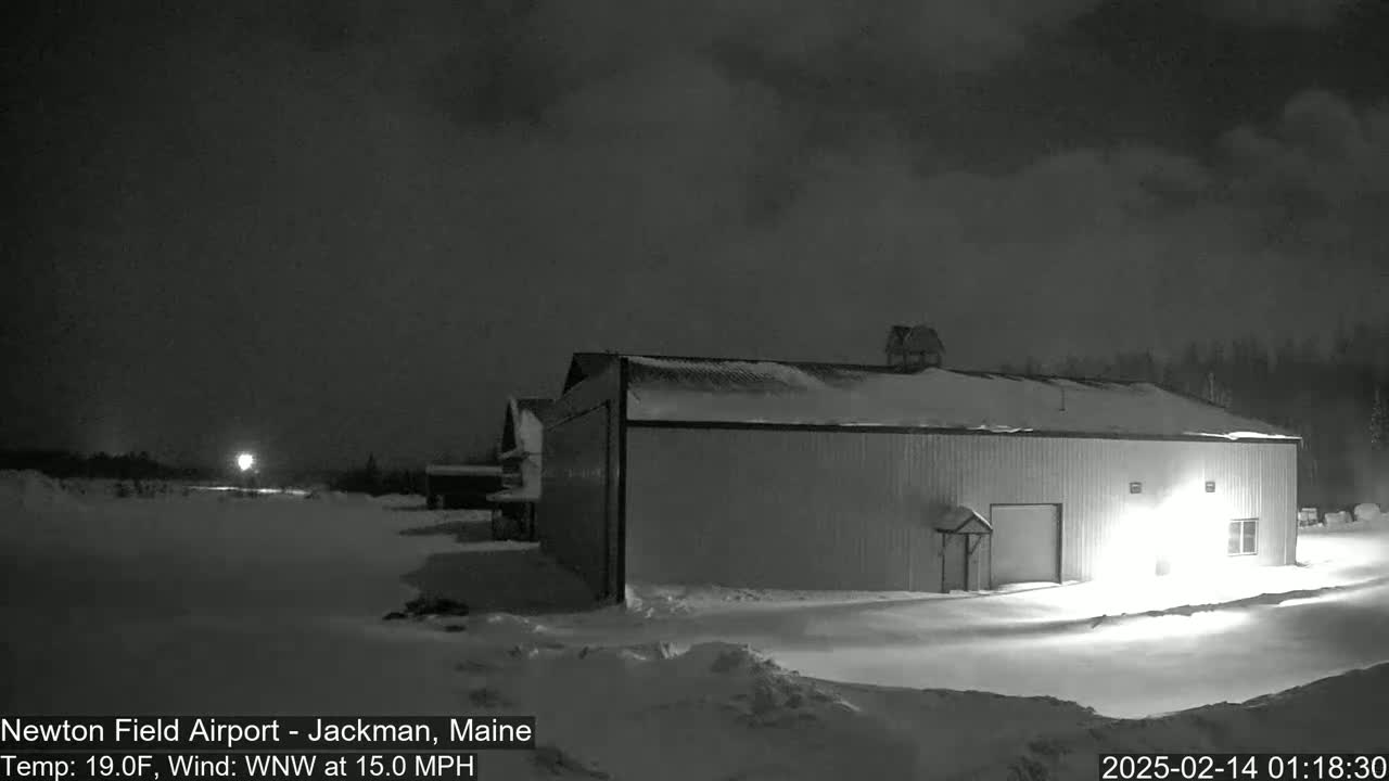 A large, snow-covered building sits in a snow-covered field at night under a dark, cloudy sky with a wind of 15 mph.