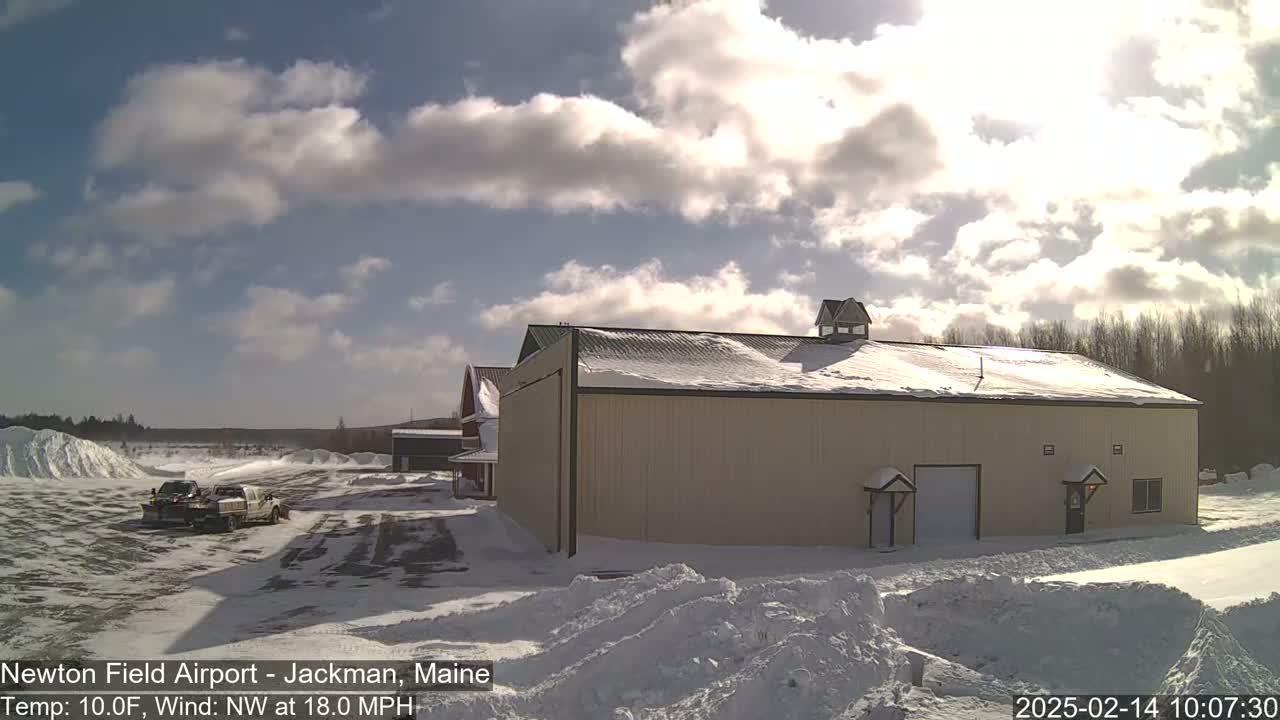 A large tan building with snow on its roof sits in a snowy landscape under a partly sunny sky, with two snowplows parked nearby.