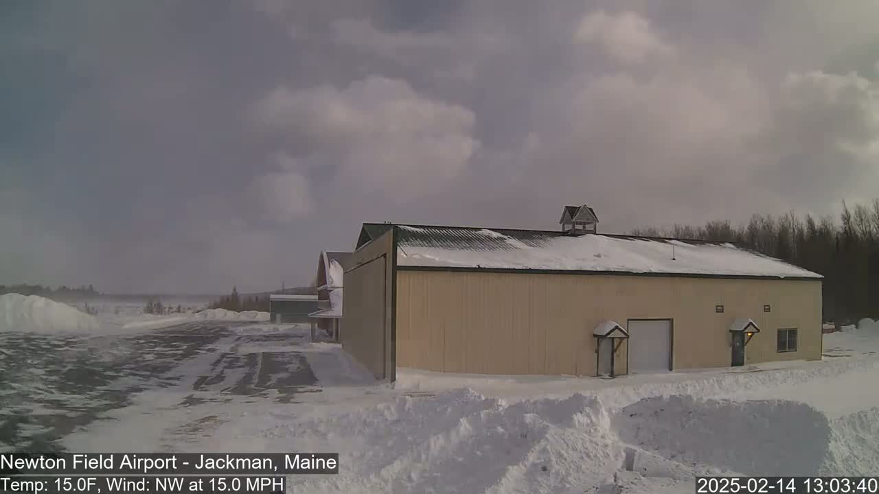 A large tan building with snow on its roof sits on a snowy airfield under a cloudy sky with a northwest wind.