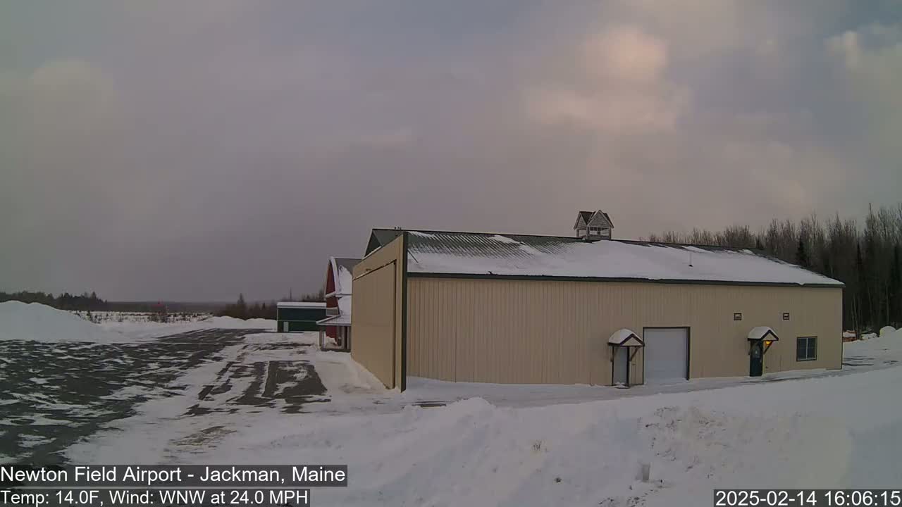 A large tan hangar sits on a snow-covered airfield under a cloudy sky with a 24 mph wind.