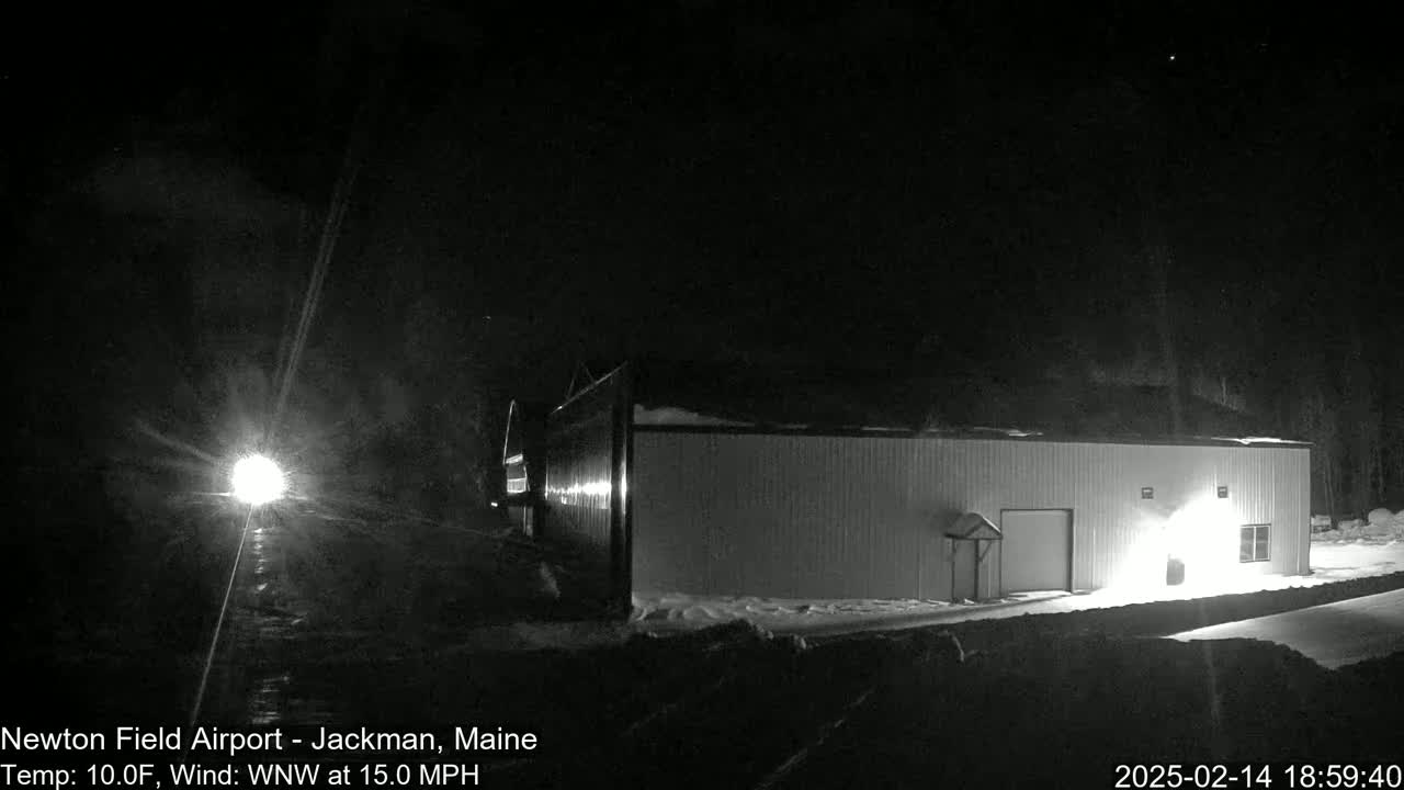 A long, light-colored building is illuminated at night in snowy conditions, with a bright light source visible in the distance and a WNW wind at 15 mph.