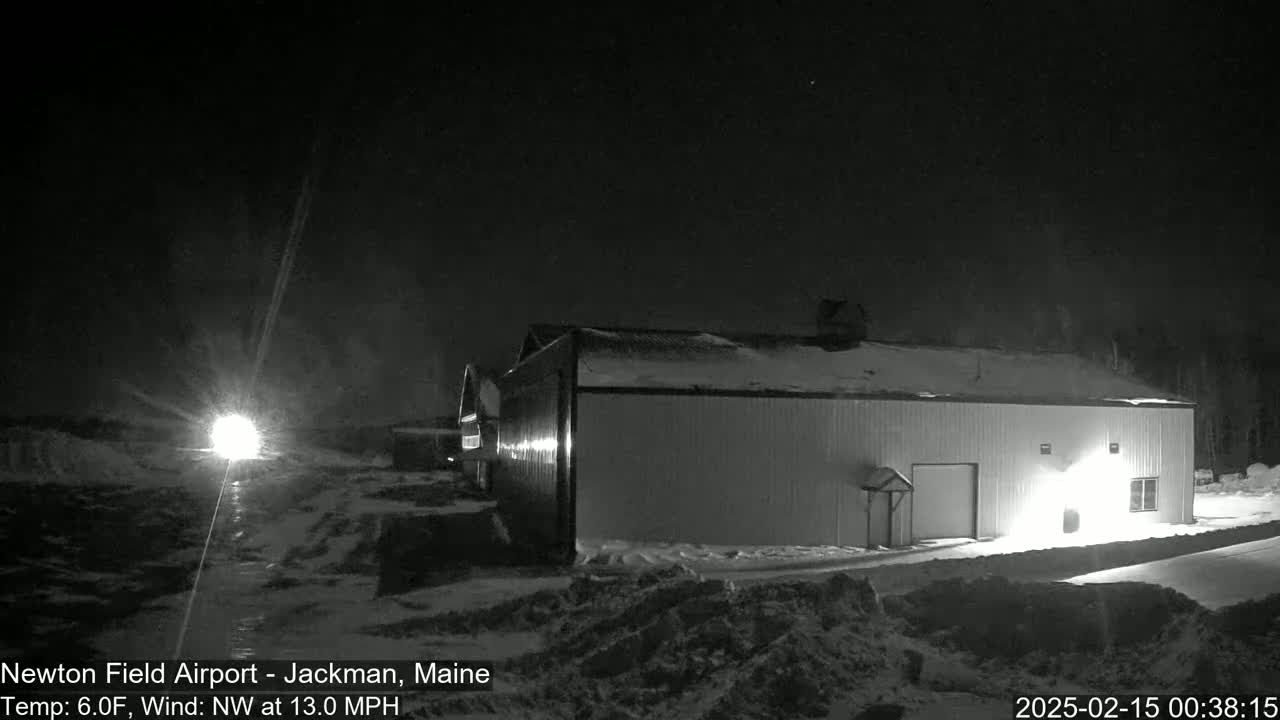A snow-covered airfield at night, illuminated by headlights in the distance and a building's exterior light, with a northwest wind at 13 mph and a temperature of 6°F.