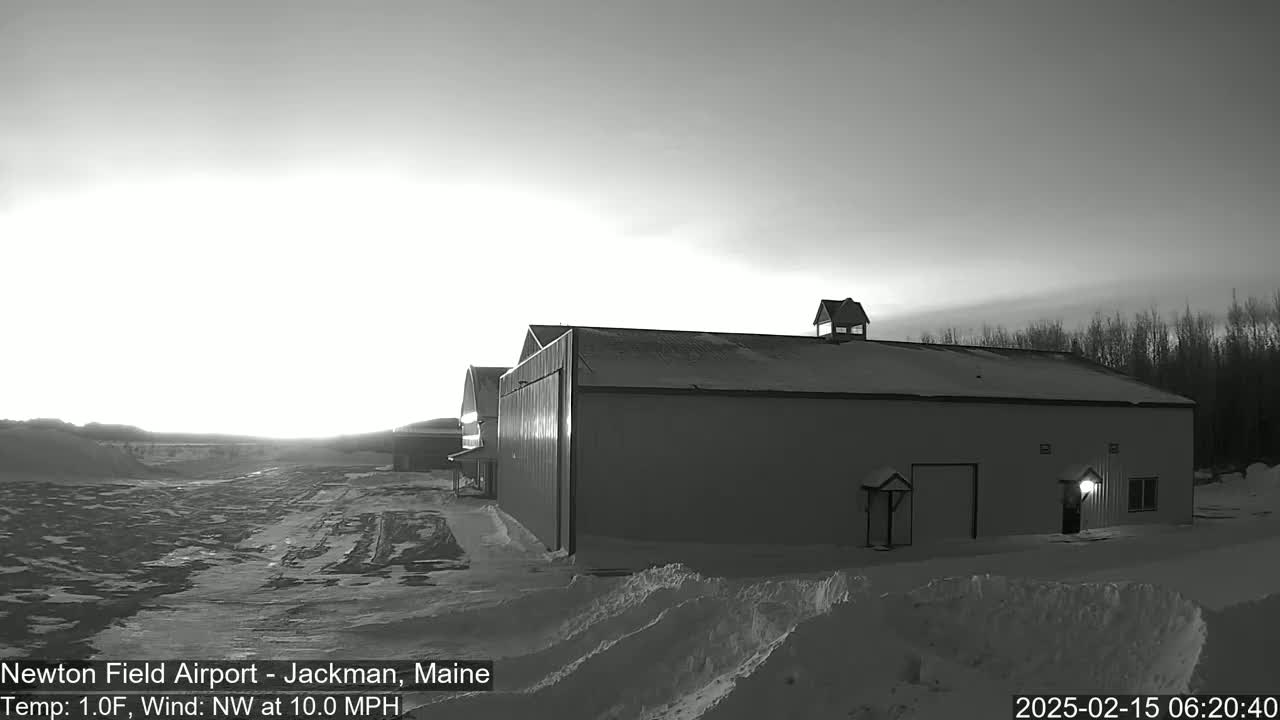 A large hangar-like building sits in a snow-covered airfield at sunrise, under a mostly clear sky with a northwest wind.