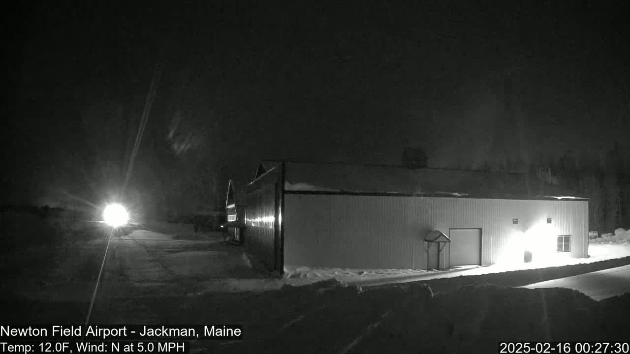 A snow-covered hangar is illuminated at night under a dark, clear sky with a light source visible in the distance.