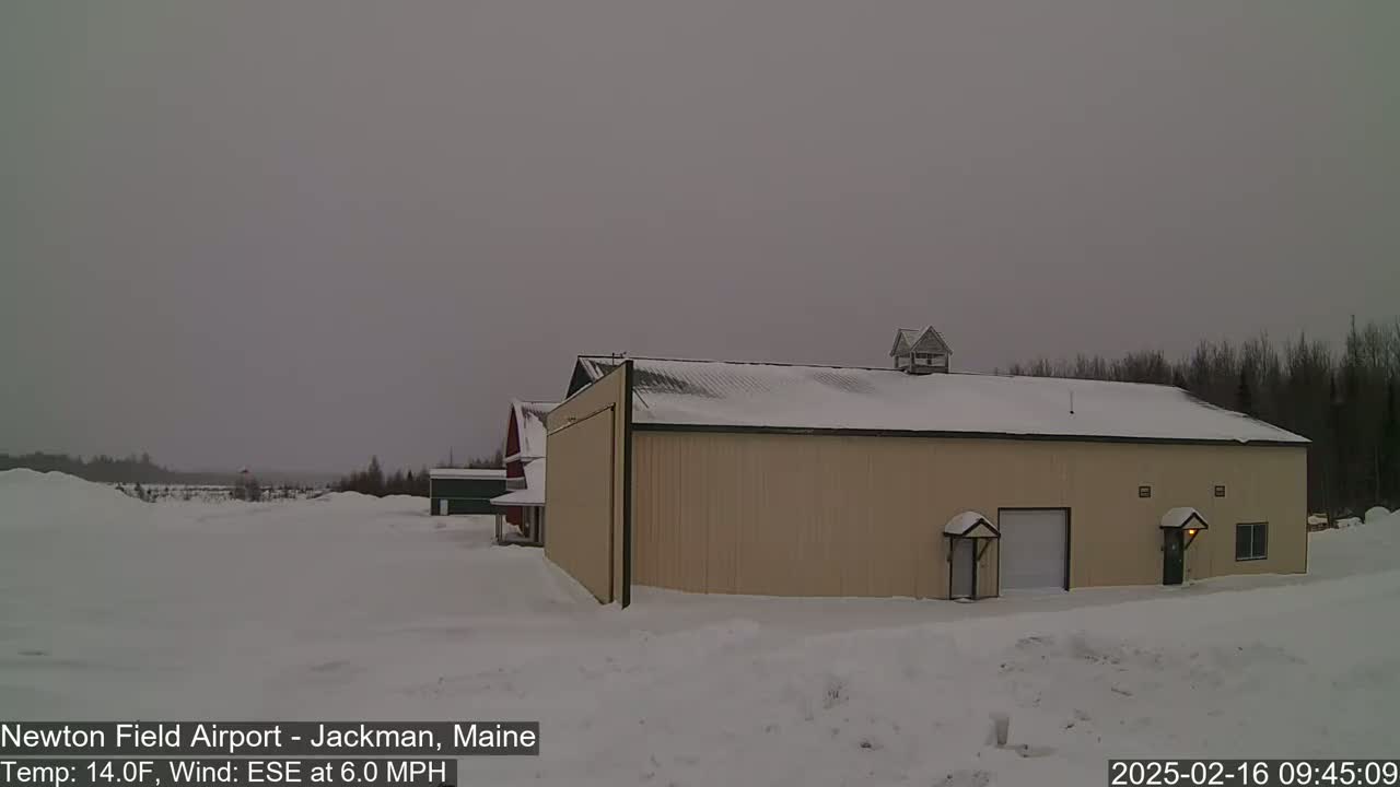 A tan building with a snow-covered roof and a smaller red building behind it sit in a snowy landscape under an overcast sky.