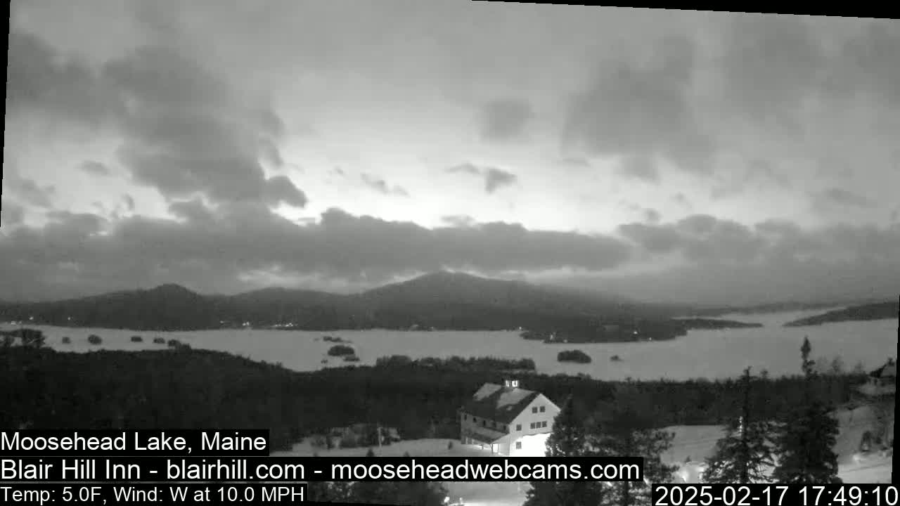 A grayscale view of a lake and distant mountains under a cloudy sky, with a building visible in the foreground.