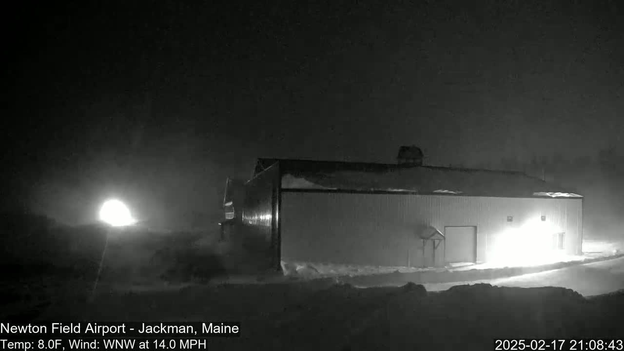 A large, snow-covered building is illuminated at night in snowy conditions with a 14 mph wind.