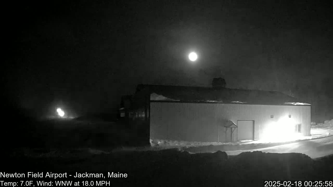 A bright moon illuminates a snow-covered landscape at night, with a large building and a bright light source visible in the foreground, under a WNW wind of 18 mph at 7°F.