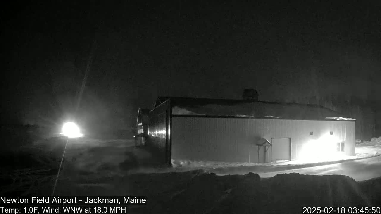 A snow-covered hangar is illuminated at night by a bright light source, with a strong wind blowing.