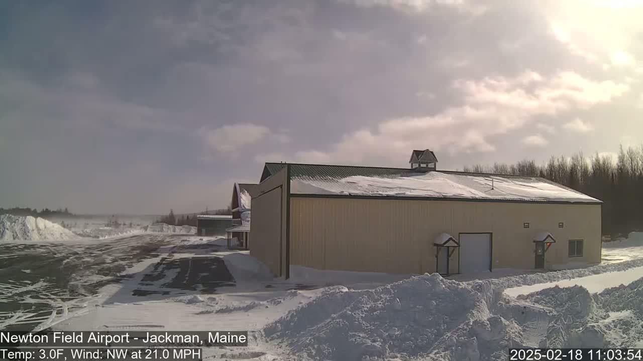 A large, tan building with a snow-covered roof sits on a snowy airfield under a partly cloudy sky, with a northwest wind blowing at 21 mph.