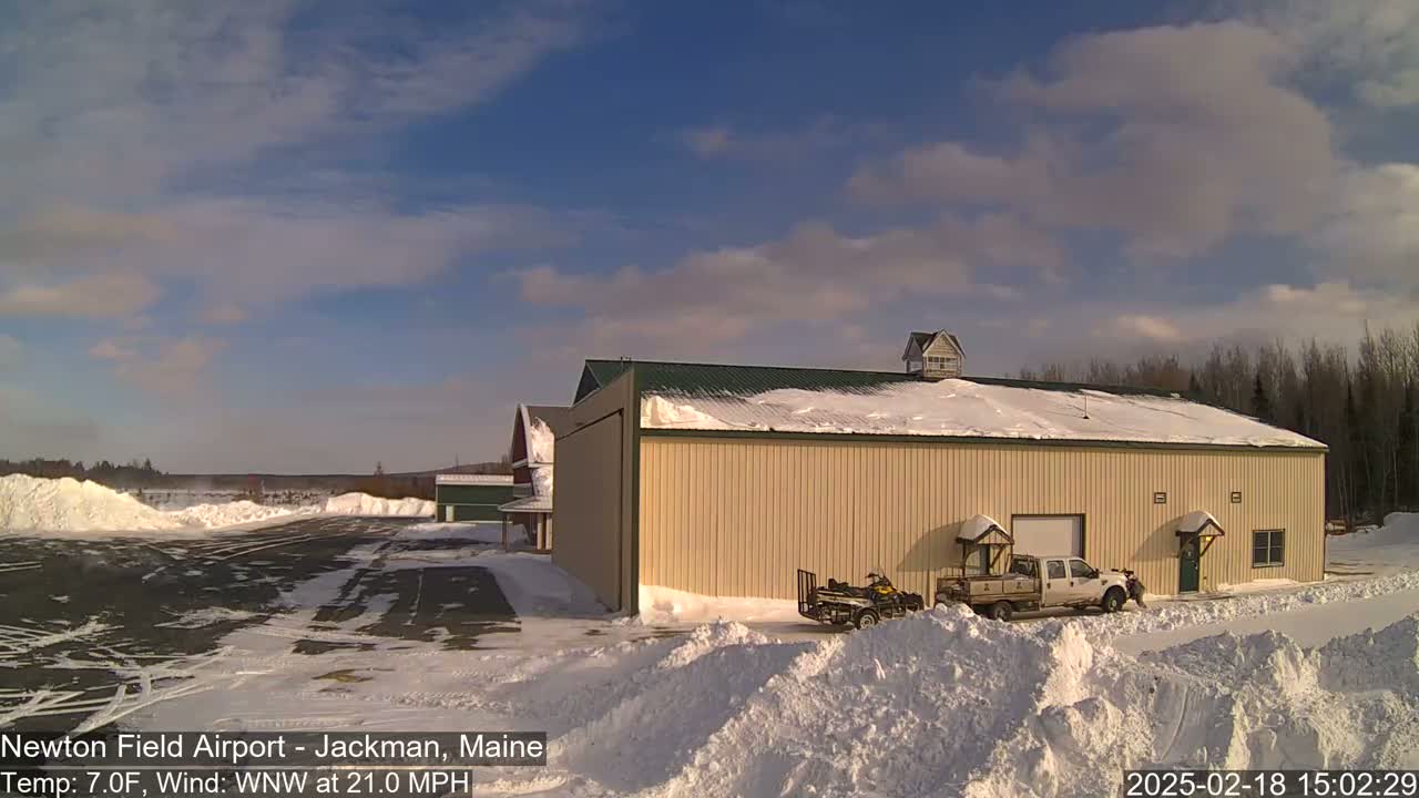A large tan hangar sits in a snowy airfield under a partly cloudy blue sky.