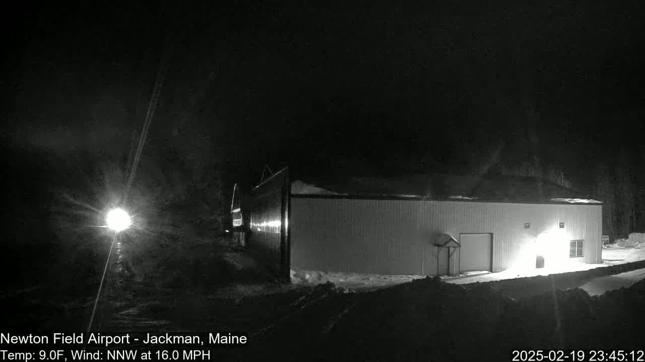 A long, white building is illuminated at night in snowy conditions under a dark sky with a northwest wind.