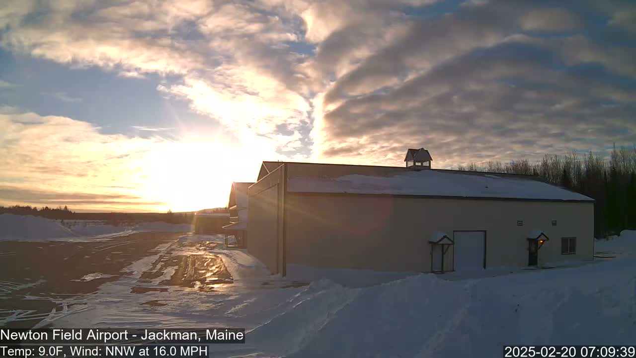A large tan building with a snowy roof sits in a snow-covered airfield at sunset under a partly cloudy sky.