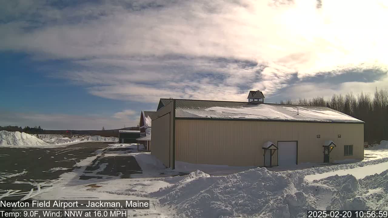 A large tan building with a snow-covered roof sits in a snowy airfield under a partly sunny sky with a brisk north-northwest wind.
