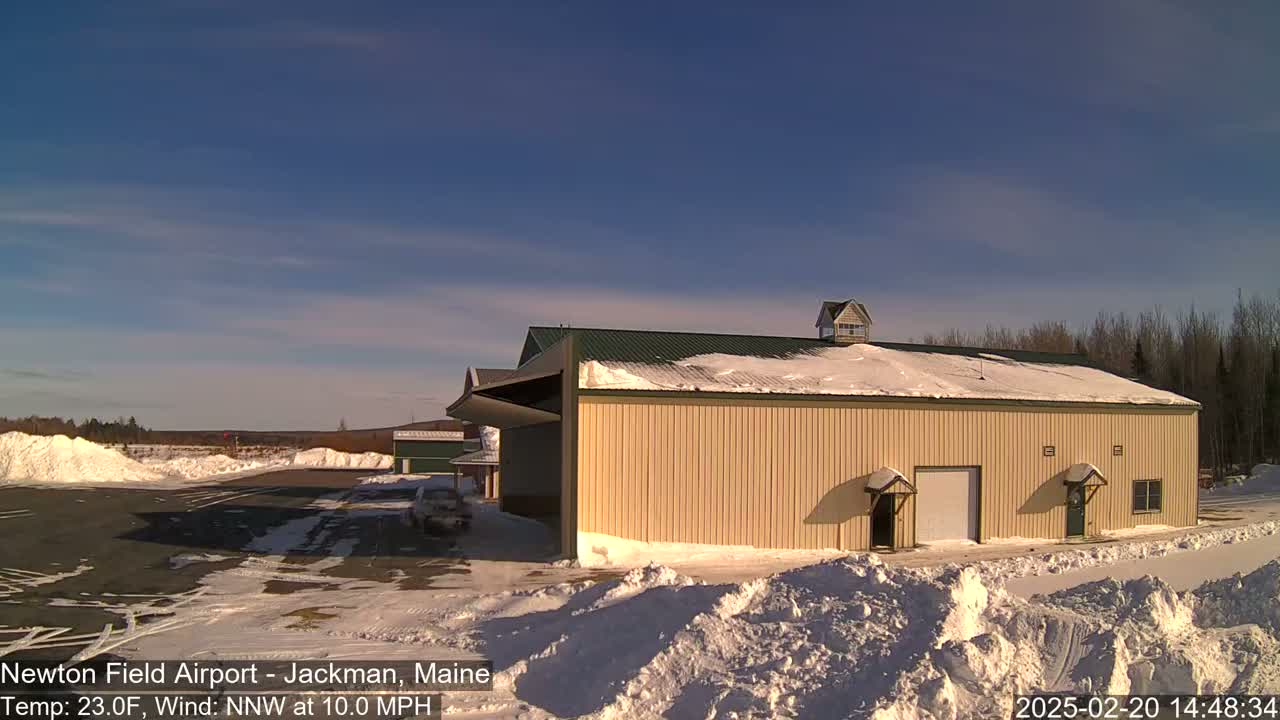 A large tan hangar with a snow-covered roof sits on a snowy airfield under a clear blue sky.