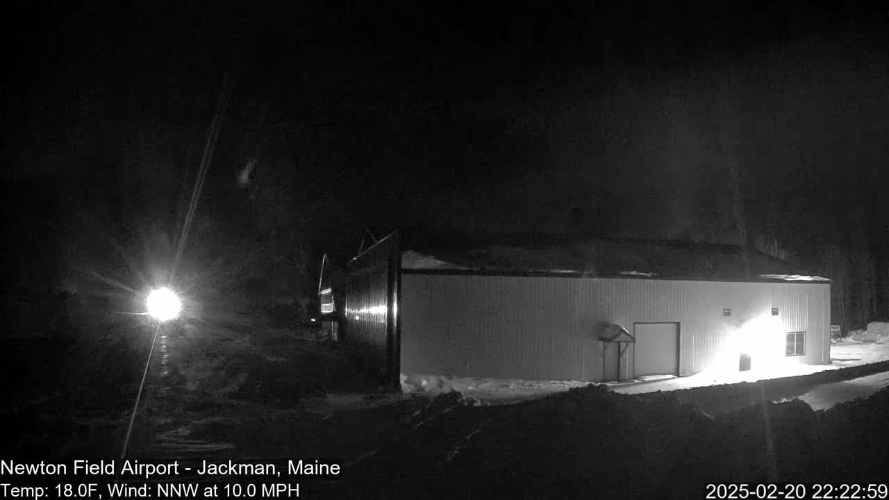 A long, light-colored building is partially illuminated at night in snowy conditions with a bright light source visible in the distance.