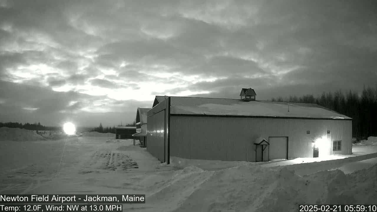 A large hangar sits in a snowy landscape under a cloudy, bright sky before sunrise.