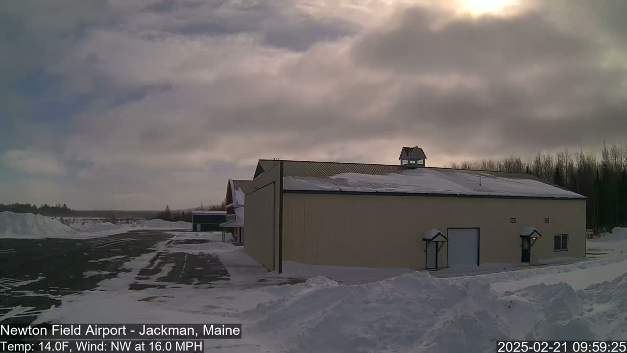 A large tan building with snow on its roof sits in a snowy landscape under a cloudy sky.