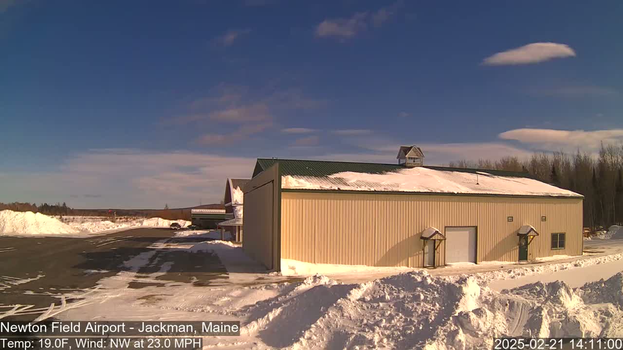 A large tan building with a snow-covered green roof sits on a snow-covered airfield under a mostly clear blue sky with a few wispy clouds; a strong northwest wind is blowing.