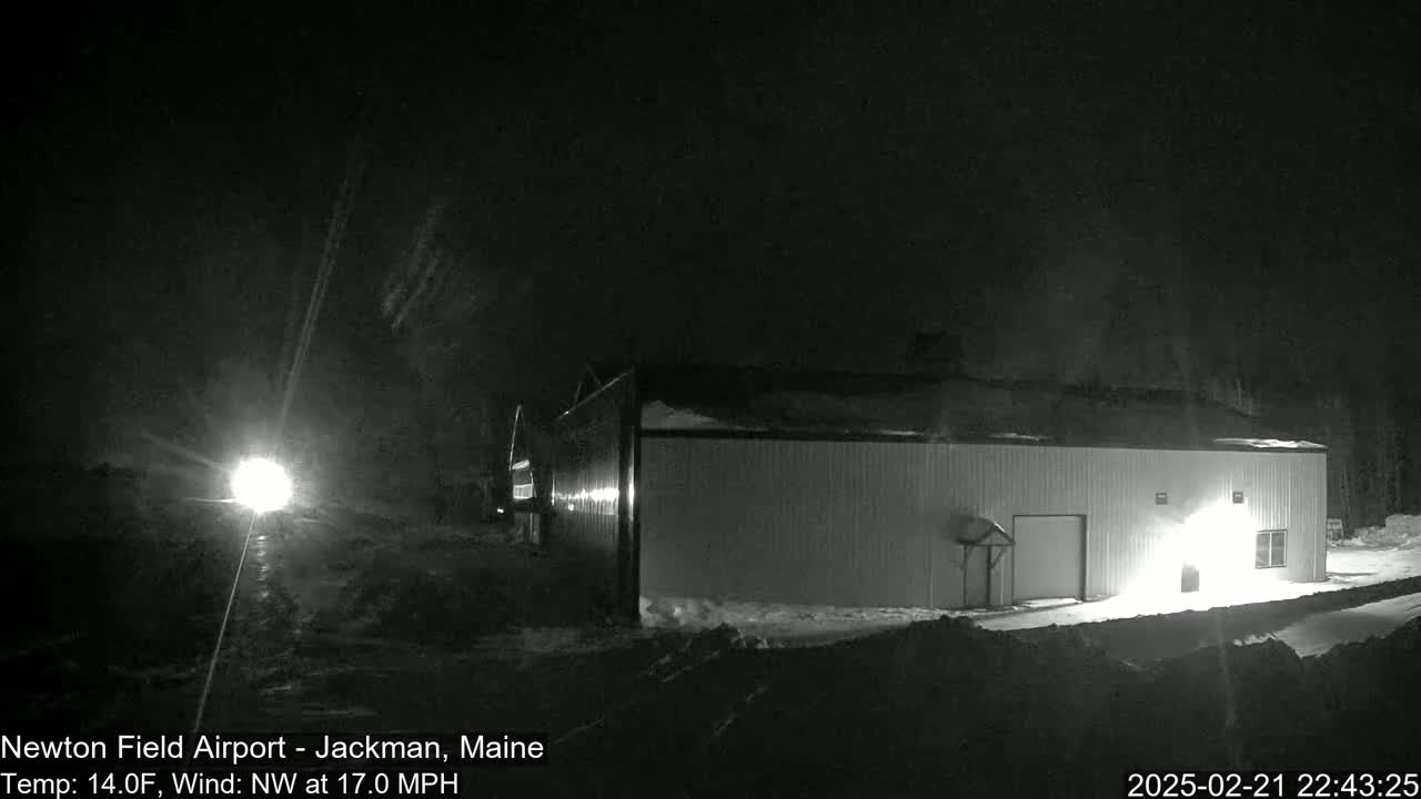 A long, low building is illuminated at night in snowy conditions under a dark sky with a bright light visible in the distance.