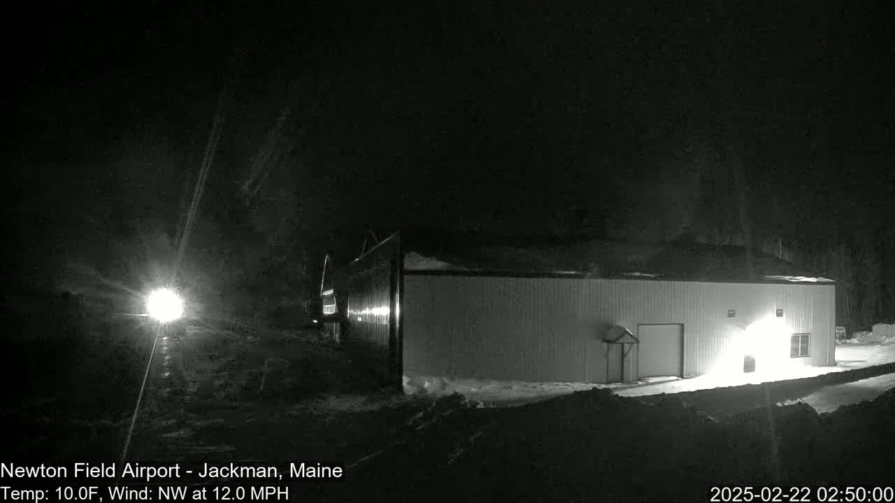 A snow-covered, light-colored building is illuminated at night during a snow flurry with a northwest wind.