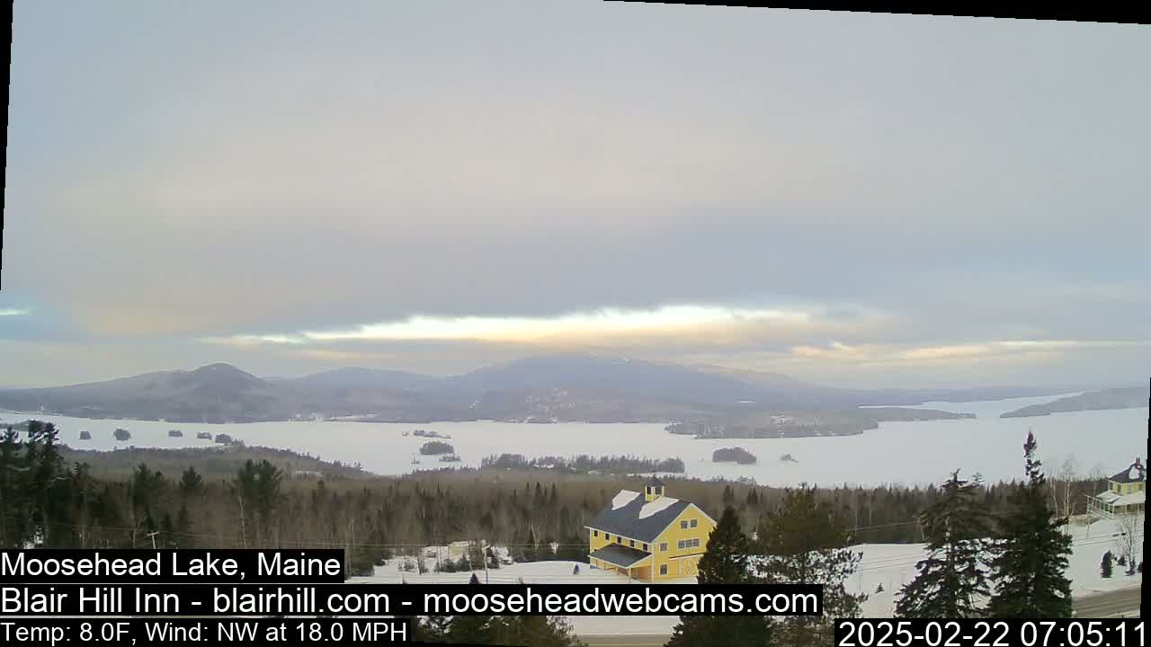 A partially frozen lake stretches to snow-covered mountains under a mostly cloudy, light sky with a northwest wind blowing at 18 mph.