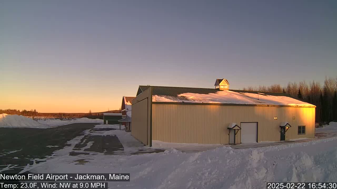 A large tan hangar sits on a snow-covered airfield at sunset under a clear sky.
