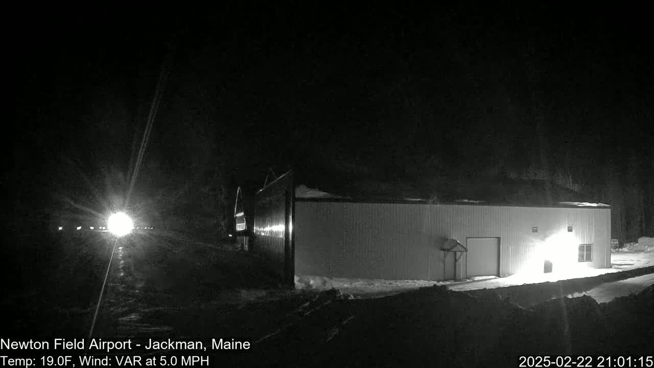 A long, low building is illuminated at night in snowy conditions with a vehicle's headlights visible in the distance.