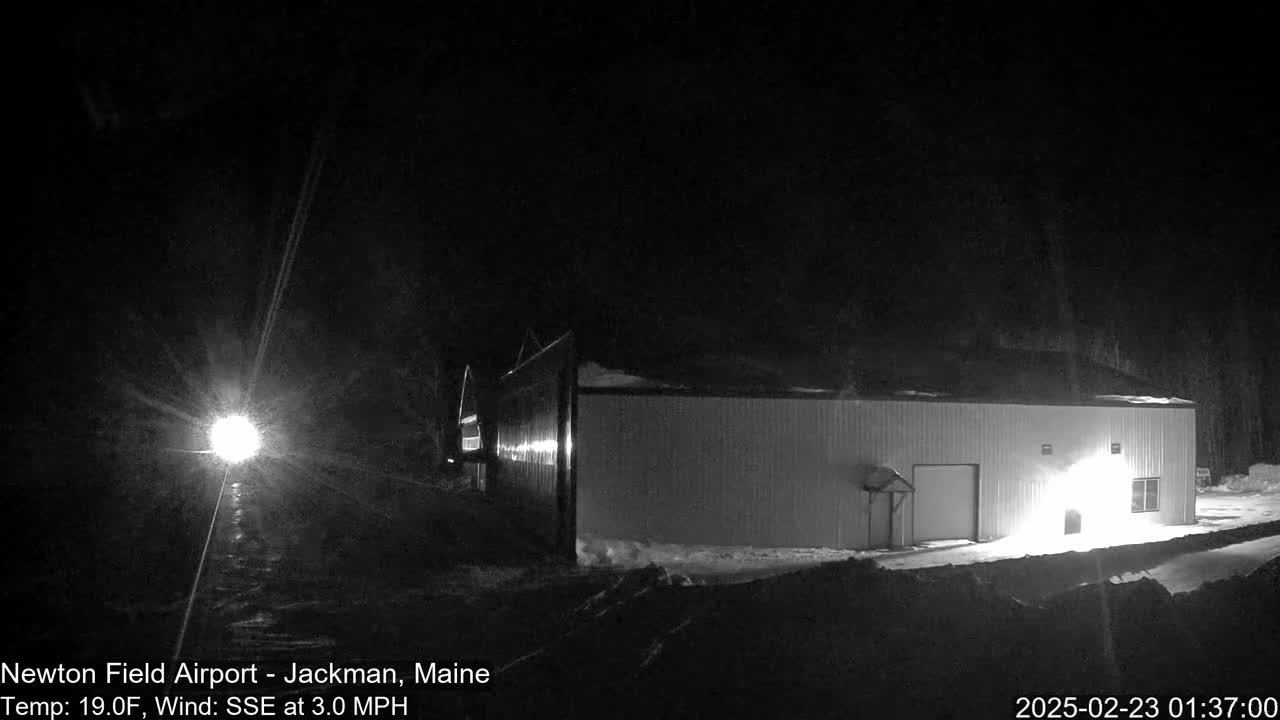 A long, light-colored building is visible at night in snowy conditions with a bright light source in the distance and a light wind.