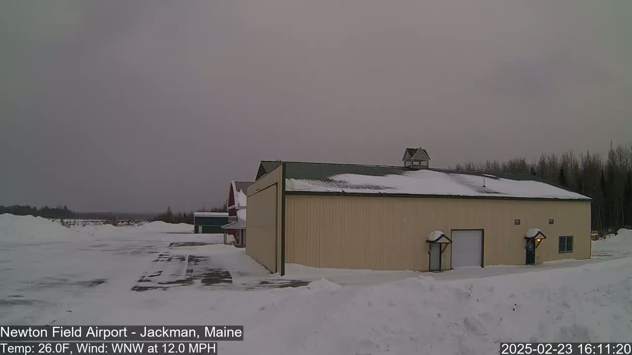 A large tan building with a hangar door sits in a snowy airfield under an overcast sky.