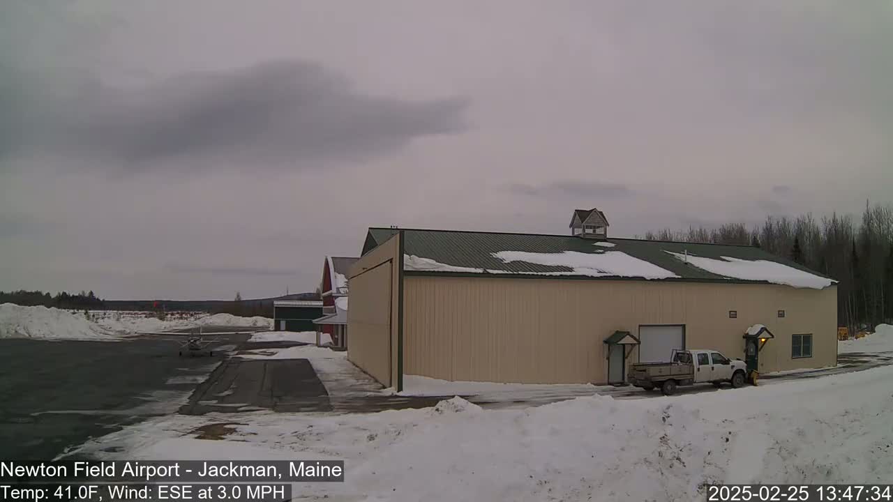 A tan hangar sits on a snow-covered airfield under an overcast sky, with a small airplane and a pickup truck nearby.