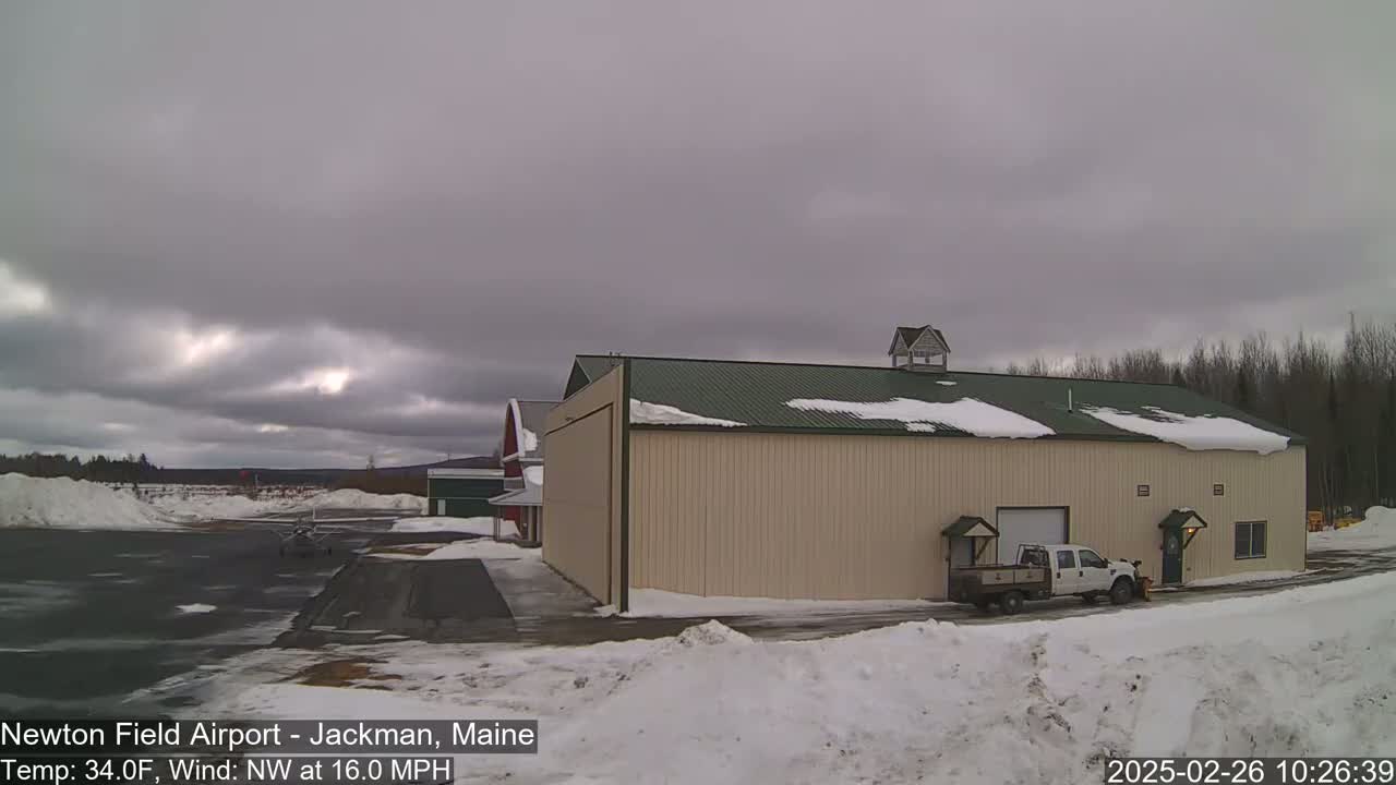 A tan hangar with a green roof sits on a snow-covered airfield under an overcast sky, with a pickup truck parked in front.