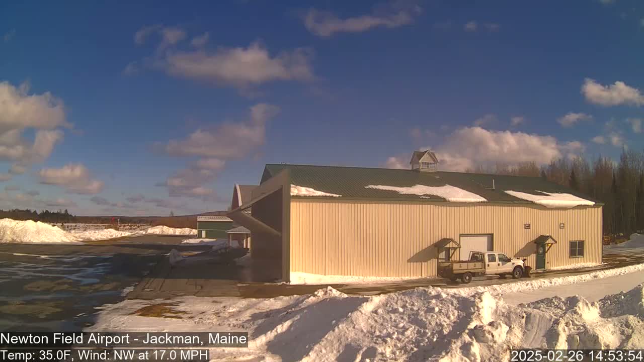 A large tan hangar with a green roof sits on a snowy airfield under a partly cloudy blue sky.