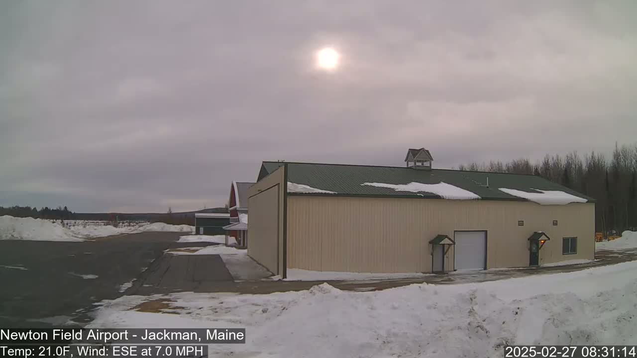 A tan hangar and a red barn sit on a snowy airfield under an overcast sky with a faint sun visible.
