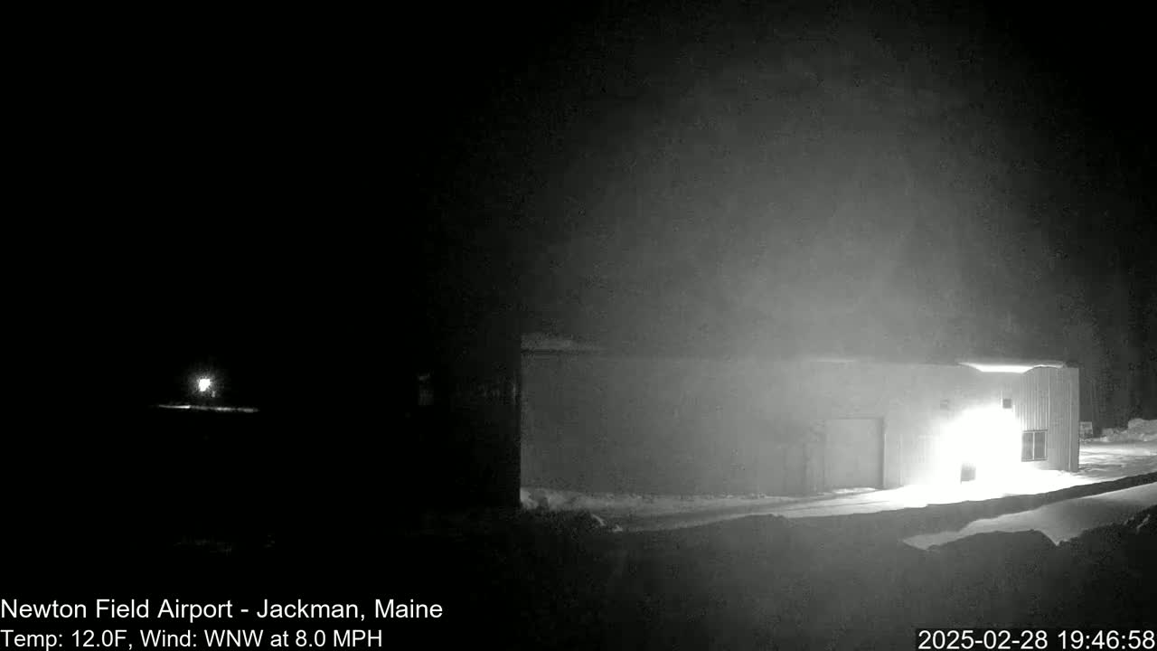 A brightly lit building is visible at night in snowy conditions with a faint light in the distance.