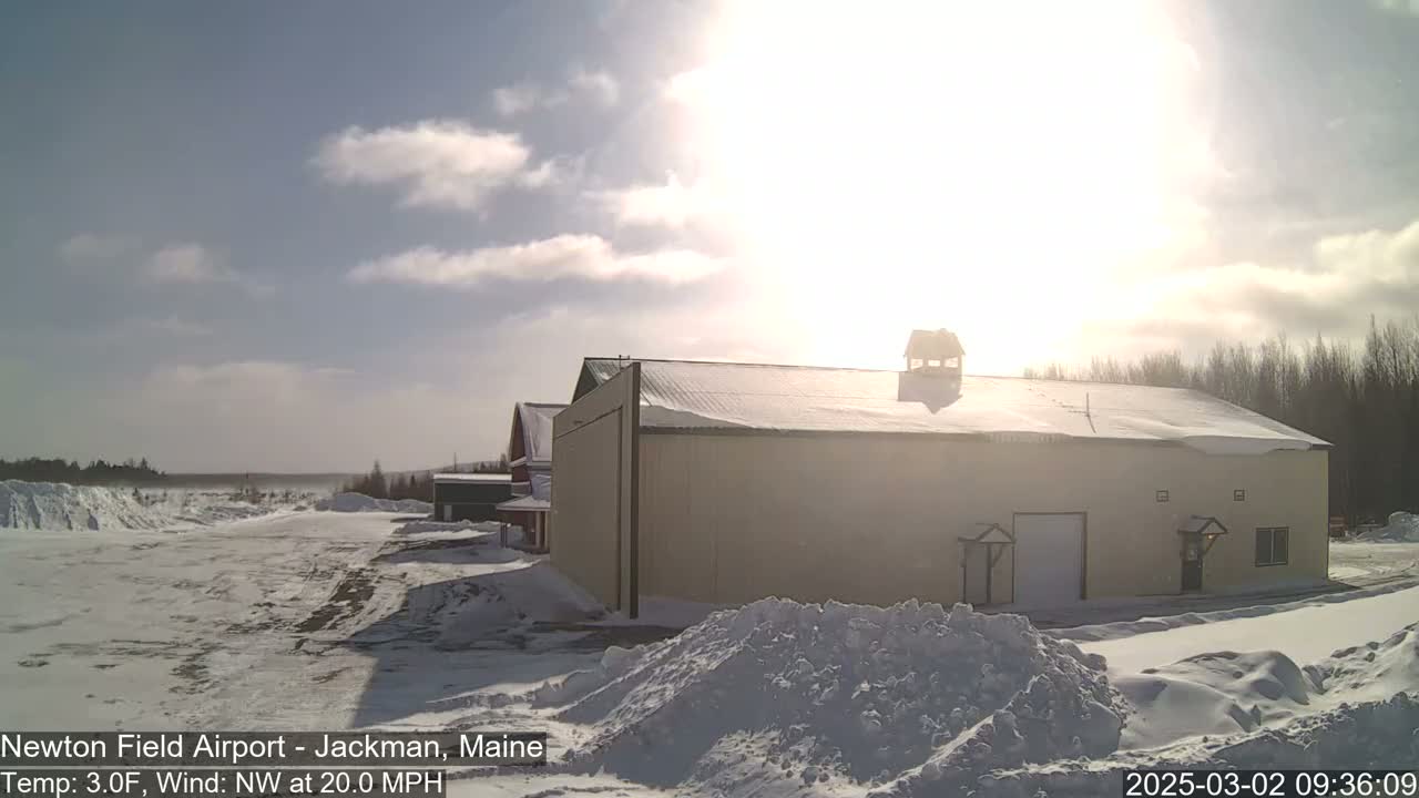 A snow-covered airfield with a large tan building and smaller structures in the background under a bright, sunny sky with some clouds and a northwest wind.