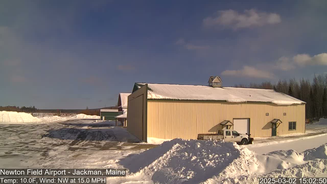 A large tan building with a snow-covered roof sits in a snowy airfield under a mostly sunny sky.