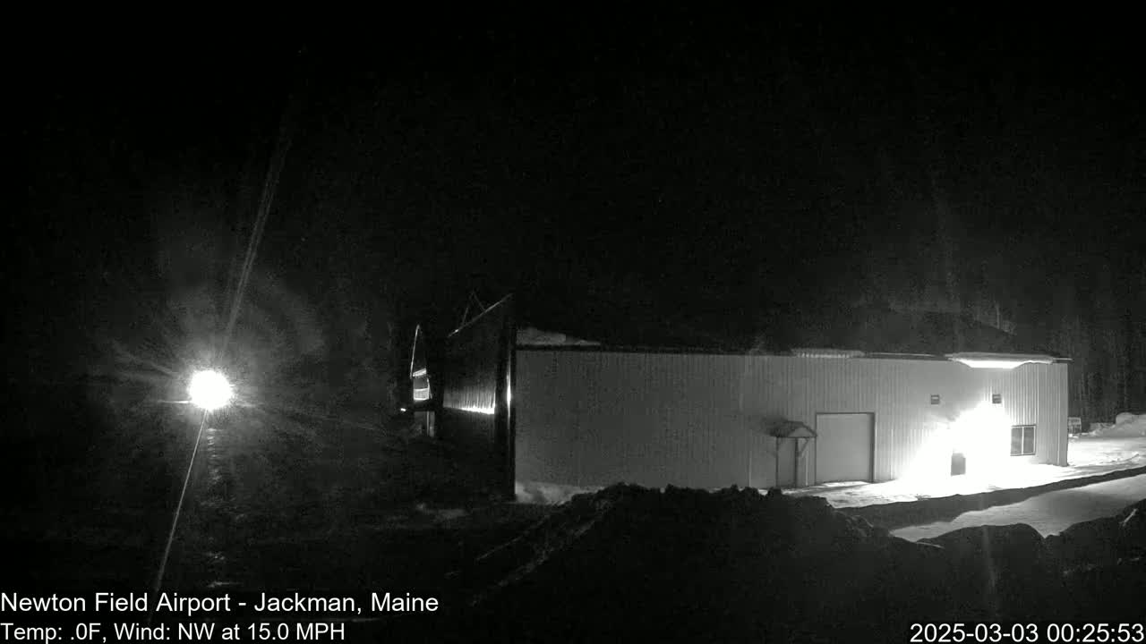 A brightly lit, long, rectangular building is seen at night in snowy conditions with a northwest wind of 15 mph.