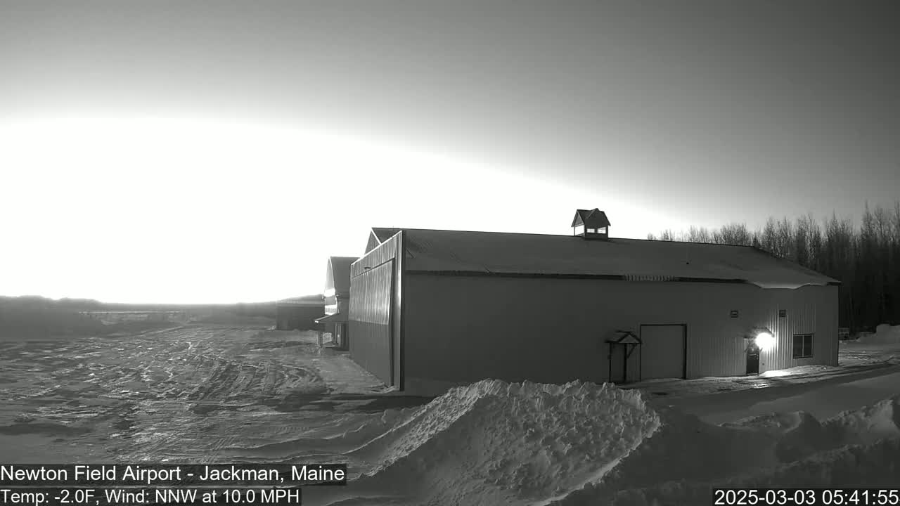 A large, snow-covered hangar sits on a snowy airfield during a bright, pre-dawn sunrise with a north-northwest wind at 10 miles per hour and a temperature of -2 degrees Fahrenheit.