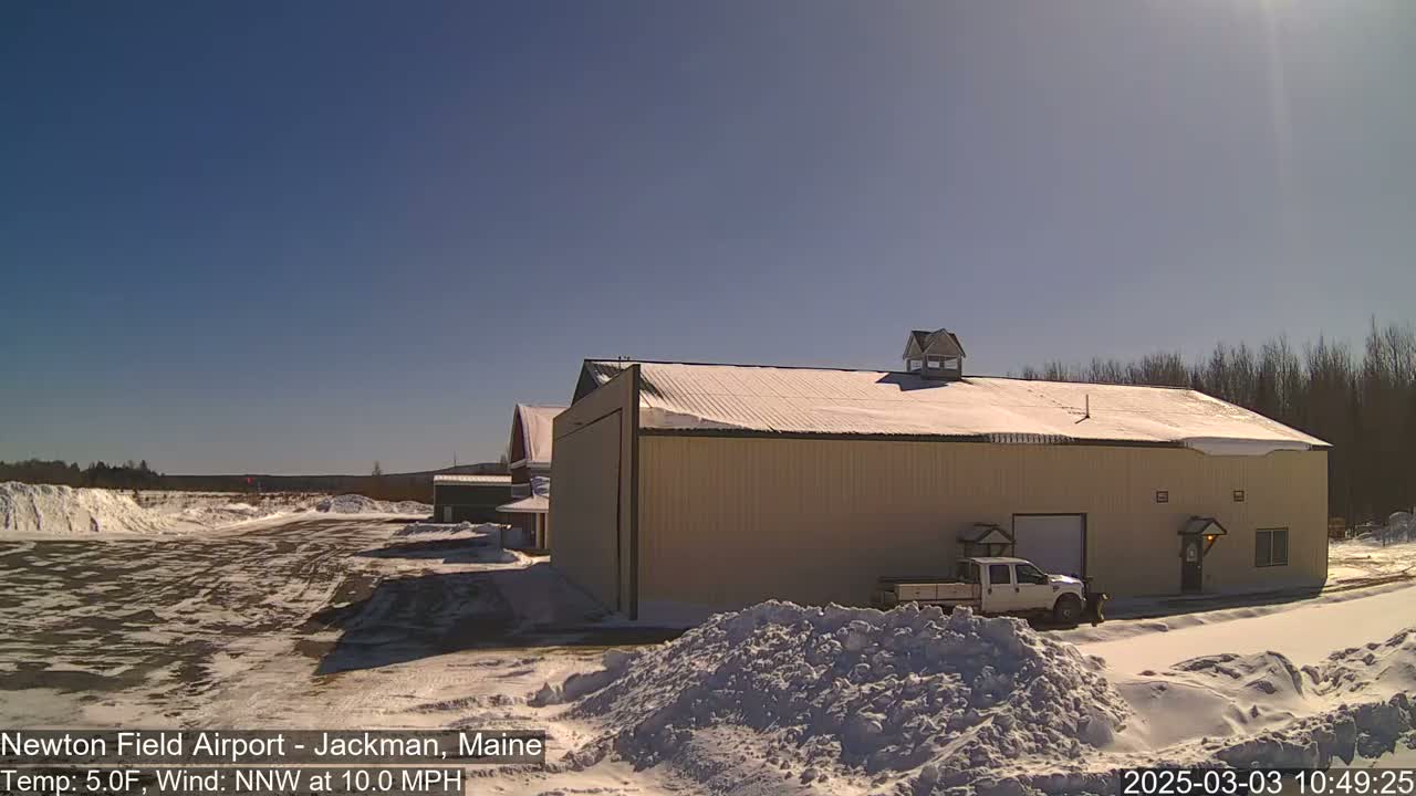 A large tan building with a snow-covered roof sits on a snowy airfield under a clear blue sky.