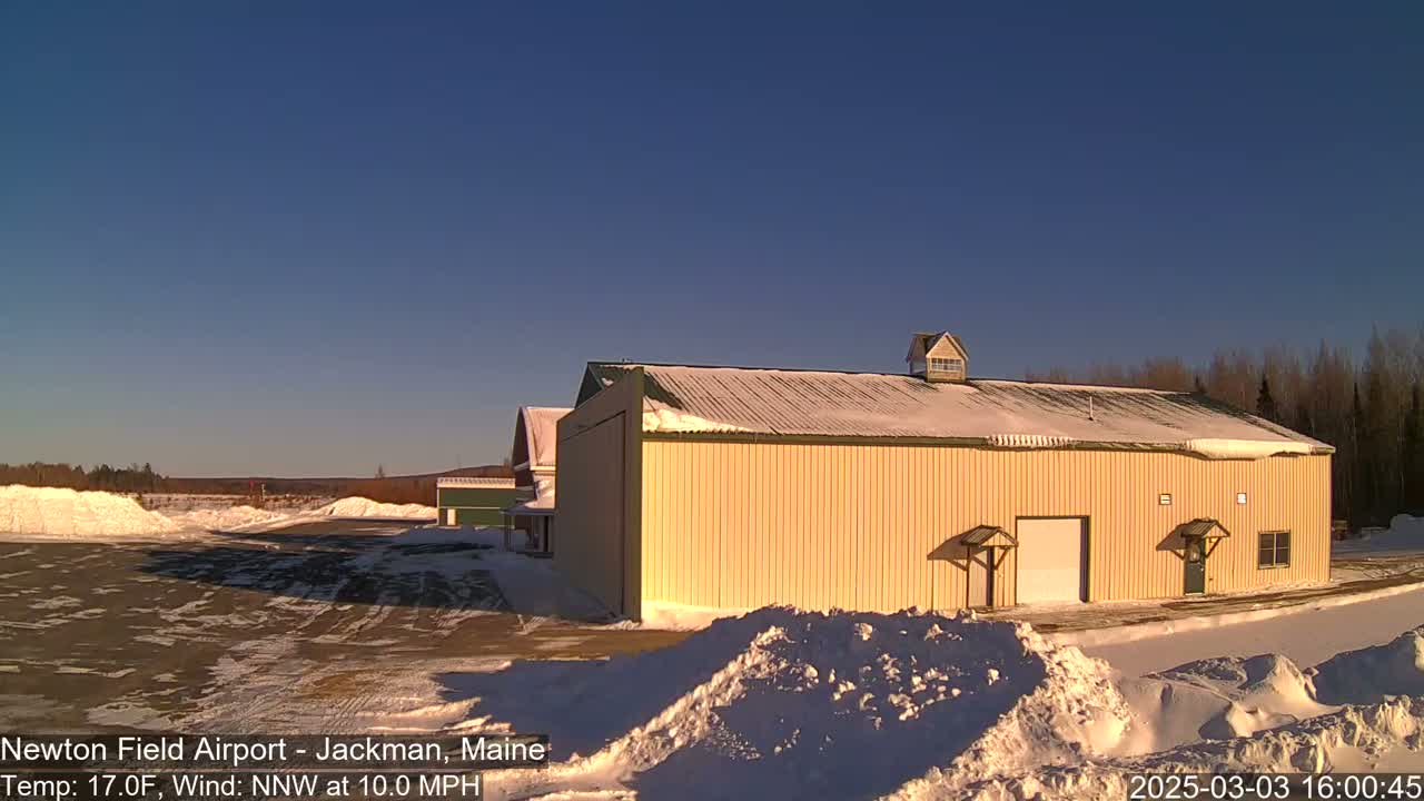 A large, light-yellow hangar sits on a snowy airfield under a clear, blue sky, with a gentle wind.