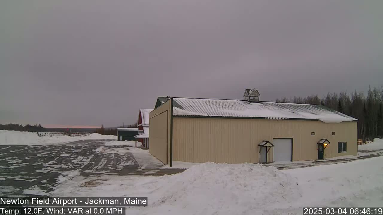 A large tan hangar sits on a snow-covered airfield under an overcast sky.